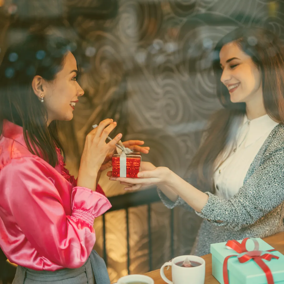 Zwei Frauen im Café, eine überreicht der anderen ein kleines rotes Geschenk mit Schleife, beide lachen