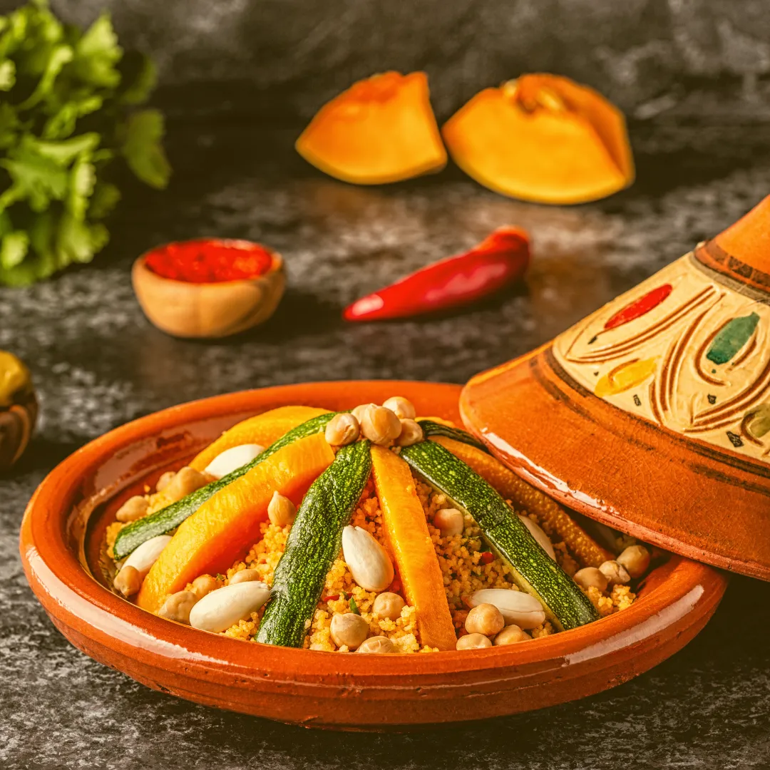 Couscous mit Gemüse in traditioneller Tonschale Couscous mit Kichererbsen, Bohnen und Gewürzen in bemalter Tonschale, daneben Chili und Kräuter