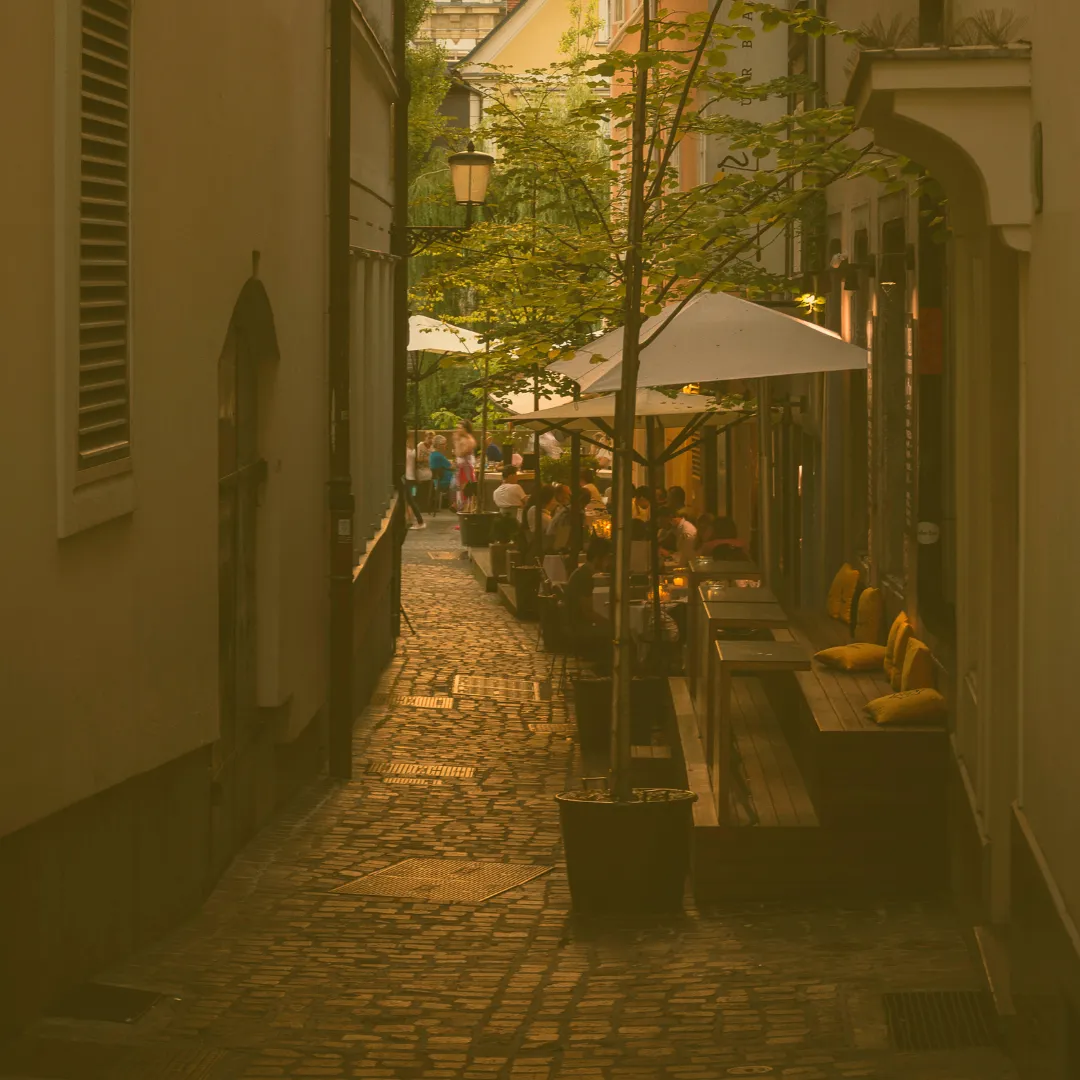 Schmale Altstadtgasse mit Kopfsteinpflaster, Café-Tischen und Sonnenschirmen bei warmem Licht