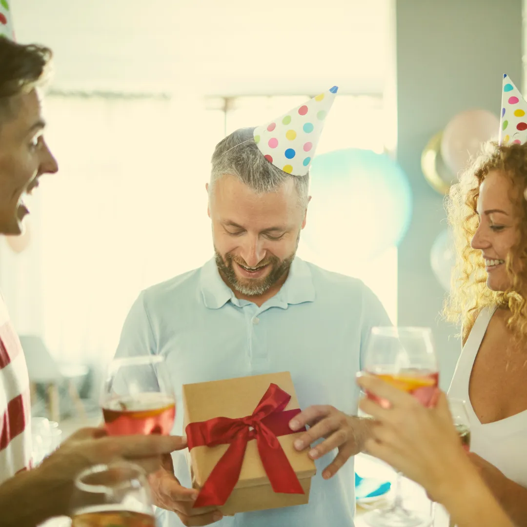 Mann mit Partyhut erhält ein Geschenk mit roter Schleife bei einer Geburtstagsfeier im Freundeskreis